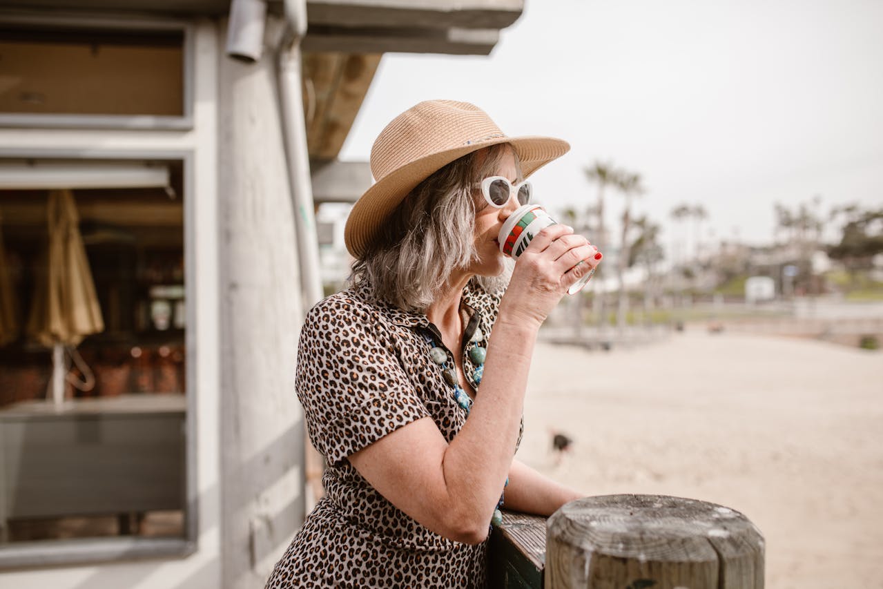 Woman in a leopard print dress and hat drinking from a coffee cup on the beach.