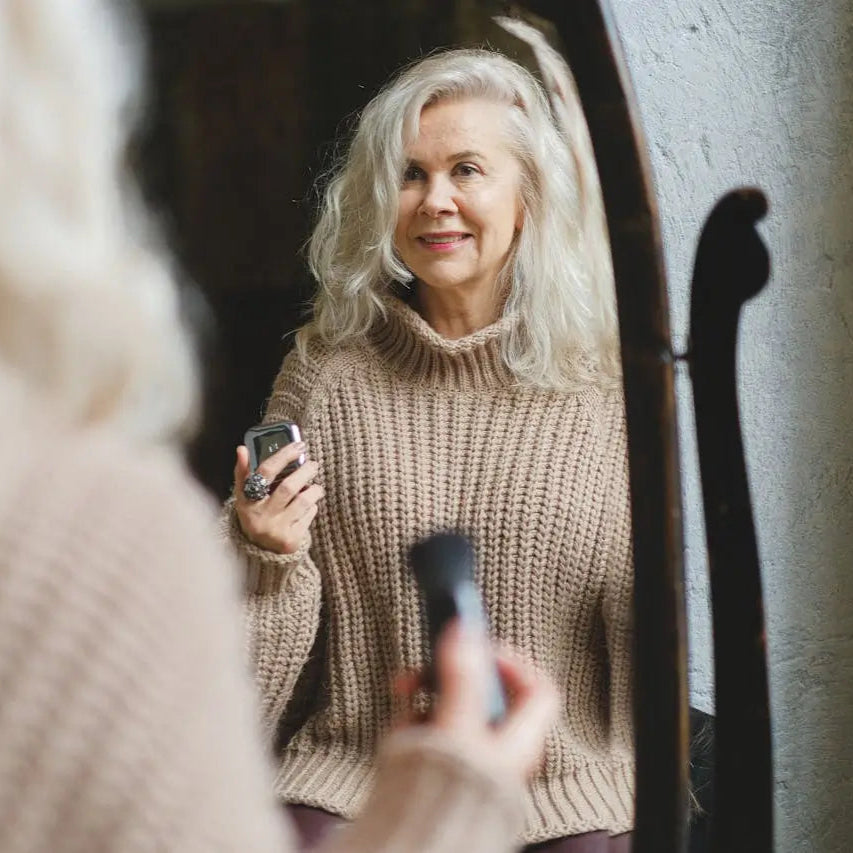 And older white woman with long greyish white hair smiles at herself in a mirror, holding a makeup brush and compact